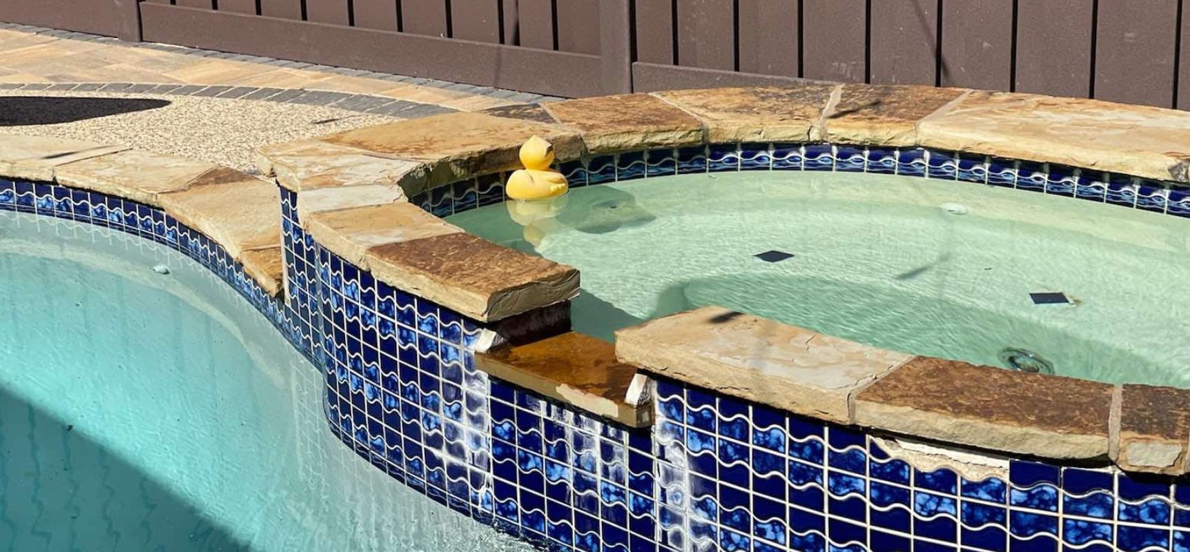 A small hot tub with blue tile borders overflows into a swimming pool. A yellow rubber duck floats in the hot tub.