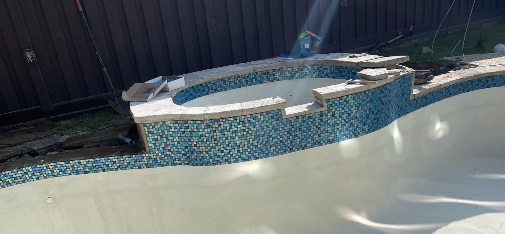 An empty swimming pool with a raised, unfinished spa area, mosaic tile siding, and construction materials on the edge.